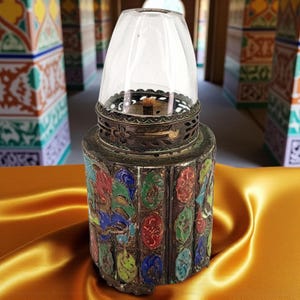 Könnte beinhalten: Eine antike Öllampe mit einem transparenten Glasschirm und einem dekorativen Metallsockel. Der Sockel weist farbenfrohe Emaille-Paneele mit floralen und geometrischen Mustern auf. Die Lampe steht auf einer goldenen Satin-Oberfläche.