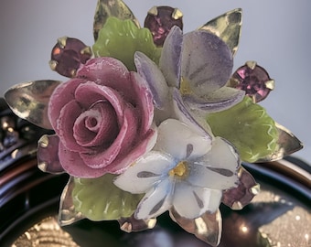 Puede incluir: Un broche vintage con un grupo de flores de porcelana con pétalos rosas, blancos y morados. Las flores están adornadas con brillantes diamantes de imitación rosas.