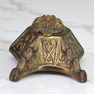 May include: Antique brass inkwell with a detailed floral lid and ornate, geometric body. The inkwell has a square base with curved feet and decorative scrollwork. The aged brass finish gives it a vintage look.