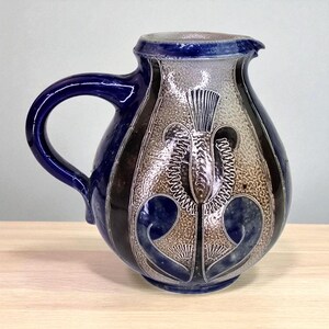 May include: A ceramic pitcher with a dark blue handle and rim. The body features a textured, light brown and silver design with dark blue and black accents. The pitcher has a floral motif and a spout for pouring.