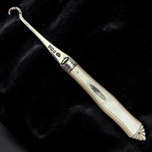 May include: A silver-toned surgical instrument with a curved hook at one end and a mother-of-pearl handle. The handle has an oval inlay and a decorative, scalloped end. The instrument is set against a dark background.