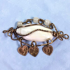 Puede incluir: Broche artesanal con una piedra blanca ovalada envuelta en alambre de cobre. Tres colgantes en forma de corazón con las letras "J", "M" y "O" cuelgan debajo. El broche está adornado con tres pequeñas cuentas transparentes.