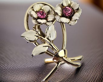 Puede incluir: Un broche de tono dorado con dos flores blancas con centros de pedrería rosa. Las flores están dispuestas en forma de ramo con hojas y tallos dorados.