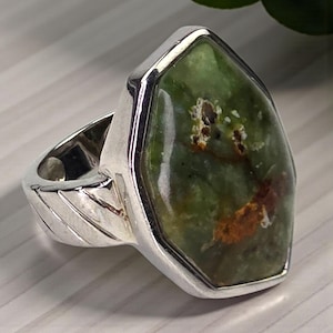 May include: A silver ring featuring a large, irregularly shaped green gemstone with brown inclusions. The gemstone is set in a polished silver bezel. The ring's band has a textured design.