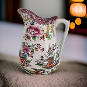 Puede incluir: Una jarra de cerámica blanca con un diseño floral rosa, verde y dorado. La jarra tiene un asa y un pico. El diseño presenta una pagoda y otros motivos de inspiración asiática.