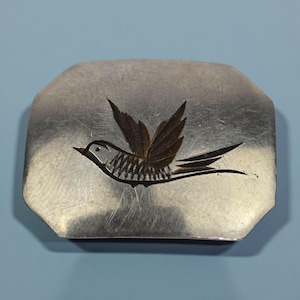 May include: A silver-coloured, rectangular trinket box featuring a detailed engraving of a bird in flight. The bird's wings are a contrasting darker colour, and the box has a slightly textured surface. The box is set against a light blue background.