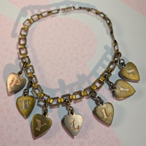 Puede incluir: Pulsera de abalorios vintage con una cadena de eslabones rectangulares y dijes en forma de corazón. Los dijes están grabados con letras en oro, plata y cobre. Una pieza de joyería única con una estética clásica y antigua.