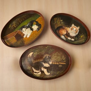 Può includere: Tre vassoi ovali con scene di gatti dipinte a mano. Ogni vassoio presenta un gatto diverso in un ambiente naturale, con toni marroni e verdi. I vassoi hanno un bordo marrone scuro e sono esposti su uno sfondo chiaro.