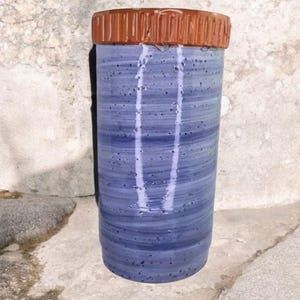 May include: A tall, cylindrical ceramic vase with a blue and white marbled design. The vase has a textured, brown rim. The vase is standing on a concrete surface.