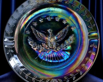 May include: Iridescent glass ashtray with a central embossed design featuring an eagle, stars, and a shield. The rim is decorated with a leaf pattern. The ashtray has a rainbow sheen, reflecting colors of blue, green, and gold.