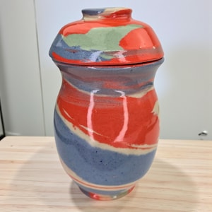 Puede incluir: Un tarro de cerámica hecho a mano con tapa. El tarro está pintado con un remolino de colores rojo, azul y verde. El tarro está sobre una superficie de madera.