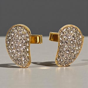 May include: A pair of gold-toned cufflinks, each with a teardrop shape. The surface of each cufflink is covered in small, sparkling, clear crystals. The cufflinks are set against a neutral gray background.