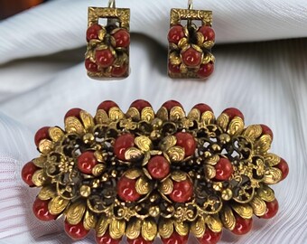 Puede incluir: Un conjunto de joyas vintage que incluye un broche y pendientes. Las piezas están hechas de metal dorado y presentan cuentas de coral rojo. El broche tiene forma ovalada y un diseño floral. Los pendientes tienen forma cuadrada y un diseño sencillo.