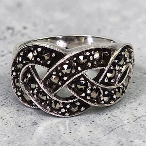 May include: A silver-toned ring with a Celtic knot design. The ring is adorned with small, dark, sparkling stones, creating a textured and detailed appearance. The ring's design features interwoven patterns, showcasing a classic and elegant style.