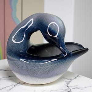 Puede incluir: Un cuenco decorativo de cerámica con forma de pato, con un degradado de tonos azules. El cuenco tiene un acabado liso y brillante y un diseño abierto único. Los detalles blancos resaltan las curvas de la forma del pato. Es un artículo decorativo.