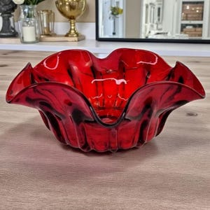 Puede incluir: Un cuenco de vidrio rojo vibrante con un borde ondulado. El cuenco tiene un color profundo y rico y un acabado brillante. El diseño recuerda a una flor, con los bordes curvándose suavemente hacia afuera. Una pieza decorativa para el hogar.