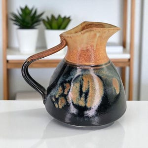 May include: A handcrafted ceramic jug with a unique design. The jug features a black base with brown and gold accents, a tan rim, and a curved handle. This decorative piece is ideal for serving beverages or as a decorative accent.