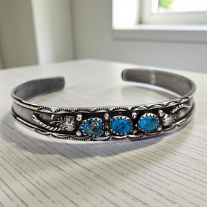 Puede incluir: Una pulsera de plata con tres piedras turquesas ovaladas. La pulsera tiene un diseño grabado con motivos de hojas y flores. El metal tiene un aspecto ligeramente envejecido.