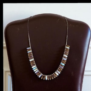 May include: A silver necklace with iridescent, teardrop-shaped beads. The beads are arranged in a graduated pattern along the necklace's curve. The necklace is displayed on a brown chair back, showcasing its design and color.