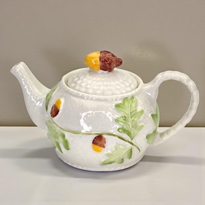 May include: White ceramic teapot with a lid and handle. The teapot is decorated with green leaves and brown and yellow acorns. The lid features a decorative acorn-shaped knob. The teapot has a spout for pouring.