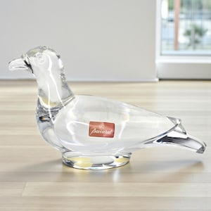 May include: A clear glass bird ornament with intricate detailing. The bird features a long beak and a rounded body. A red rectangular sticker with the word "Baccarat" is visible. The ornament is displayed on a wooden surface.