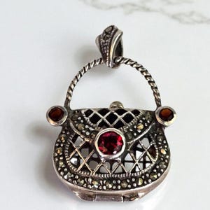 May include: A silver-toned pendant in the shape of a purse, adorned with marcasite stones and red gemstones. The purse has a detailed, openwork design with a handle and a central red gemstone. The pendant is likely vintage or antique.