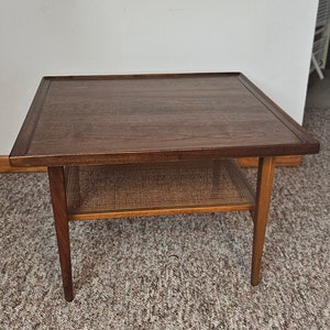 Könnte beinhalten: Ein Beistelltisch im Mid-Century-Modern-Stil mit einer dunkelbraunen Holzplatte und Beinen. Der Tisch hat ein unteres Regal mit einem gewebten Rohrdesign. Der Tisch ist ca. 61 cm breit und 61 cm lang.