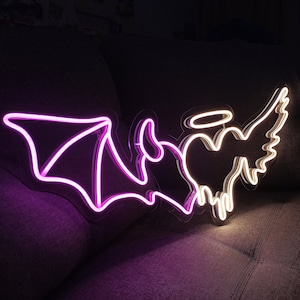 May include: Neon sign with a pink devil and a white angel with a halo and heart. The devil has wings and horns. The angel has wings and a halo. The sign is on a dark gray couch.