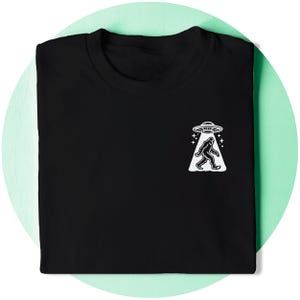 May include: Black t-shirt featuring a white graphic of a Bigfoot walking under a UFO.