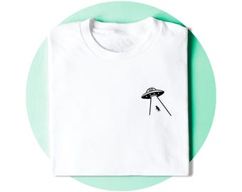 Op de afbeelding: Een wit t-shirt met ronde hals en een kleine zwarte afbeelding aan de rechterkant. Het ontwerp toont een vliegende schotel die een lichtstraal op het silhouet van een kat werpt. Het t-shirt is netjes opgevouwen op een lichtgroene cirkel.
