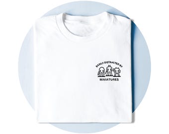 Op de afbeelding: Een wit T-shirt met korte mouwen en een zwarte afbeelding op de borst. De afbeelding toont drie miniatuurfiguren met de tekst "EASILY DISTRACTED BY" in een boog erboven en het woord "MINIATURES" in dikke zwarte letters eronder.