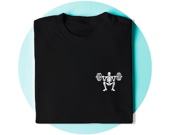 Op de afbeelding: Een zwart t-shirt met een kleine witte afbeelding aan de rechteronderkant. De afbeelding toont een skelet dat een barbell squat-oefening uitvoert, waarbij de skeletfiguur een halter met gewichten op de schouders houdt.