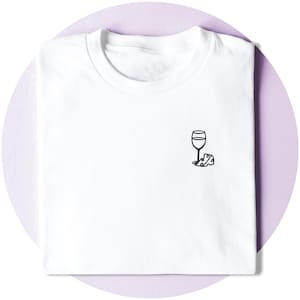 May include: White t-shirt featuring a small black illustration of a wine glass and cheese. The shirt is folded on a light purple background. The design is simple and minimalist.