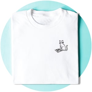 以下が含まれることがあります： 胸元に小さな黒い線画のイラストが描かれた白いTシャツ。グラフィックは、駒が並べられたチェスボードの横に座っている猫を描いています。デザインはシンプルでクリーンで、シャツの右側に配置されています。