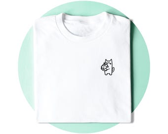 Op de afbeelding: Een wit t-shirt met een kleine, zwarte, minimalistische lijntekening op de linkerborst. De afbeelding toont een glimlachende kat die een twintigzijdige dobbelsteen vasthoudt, zoals gebruikt in rollenspellen, weergegeven in eenvoudige zwarte lijnen.