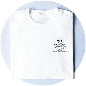 以下が含まれることがあります： 白いTシャツ。黒いイラストで、ウェイトを持ち上げる魔法使いが描かれており、その下に「LIFT, YOU FOOLS」というテキストがあります。シャツは水色の表面にきれいに折りたたまれています。