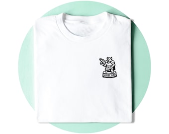 Op de afbeelding: Een wit t-shirt met ronde hals en een kleine zwarte afbeelding op de linkerborst. De afbeelding toont een gestileerd gepantserd figuur met een wapen, staand op een ronde voet met de tekst MODEL DAD duidelijk op de voorzijde gedrukt.