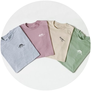 May include: Four crew neck t-shirts in light grey, dusty rose, beige, and sage green. Each shirt features a small, embroidered dinosaur skeleton graphic on the left chest area. The designs include a stegosaurus, a triceratops, and a tyrannosaurus rex.