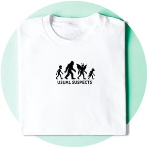 May include: White t-shirt featuring a black graphic of four cryptids, with the text "USUAL SUSPECTS" beneath. The shirt is neatly folded. The background is a light green circle.