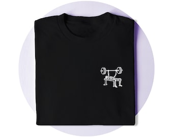 Op de afbeelding: Een zwart t-shirt met een kleine witte afbeelding op de rechterborst. De afbeelding toont een skelet dat op een gewichtsbank ligt en een bench press uitvoert met een halterstang met gewichten aan beide kanten.