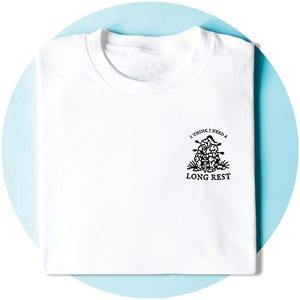 May include: White t-shirt with a crew neck. The shirt features a black graphic of a person resting with the text "I THINK I NEED A LONG REST". The shirt is folded neatly on a light blue background.