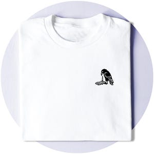 May include: A white short-sleeved t-shirt featuring a small black graphic on the chest. The graphic depicts a raven standing next to an open book with a lit candle placed on the pages. The shirt has a crew neckline and a clean, folded appearance.