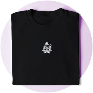 May include: Black t-shirt featuring a white graphic of a skeleton reading a book and holding a wine glass. The skeleton is seated with crossed legs. The t-shirt is folded and presented on a purple background.