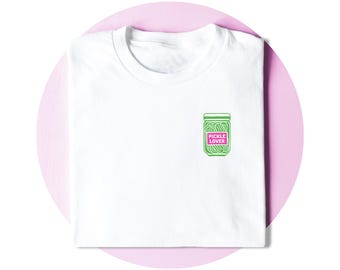 May include: A white t-shirt featuring a small graphic on the chest. The graphic depicts a green jar filled with pickles, with a pink rectangular label centered on the jar containing the white text PICKLE LOVER in all capital letters.