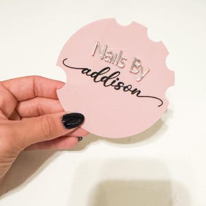 May include: A pink circle with a jagged edge, featuring the text "Nails By addison" in black cursive lettering and "Nails By" in silver lettering.
