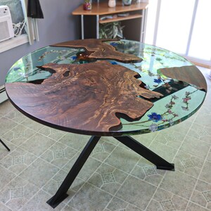 May include: A round wooden table with a live edge top and a clear epoxy resin finish. The resin contains embedded flowers and leaves. The table has a black metal base with four legs.