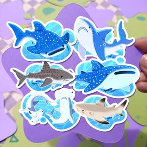 May include: A collection of colorful shark stickers. The stickers feature various shark species, including whale sharks and hammerheads, in shades of blue, gray, and tan. Each sticker is outlined and set against a blue water background.