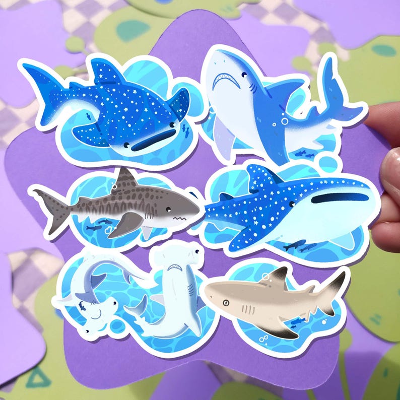 May include: A collection of colorful shark stickers. The stickers feature various shark species, including whale sharks and hammerheads, in shades of blue, gray, and tan. Each sticker is outlined and set against a blue water background.