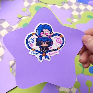 Puede incluir: Una pegatina con dos personajes de dibujos animados con pelo azul y detalles rosas, dentro de un borde en forma de corazón con las palabras "Boom!" y "Pooooow!". La pegatina está sobre un recorte en forma de estrella morada.