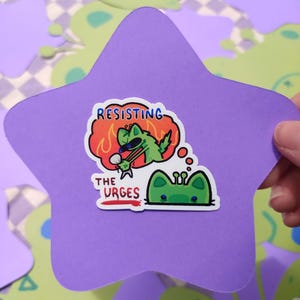 May include: A purple star-shaped sticker with a cartoon illustration. The sticker features a green cat-like character wearing sunglasses, with the text "RESISTING THE URGES" and a thought bubble with a green alien.
